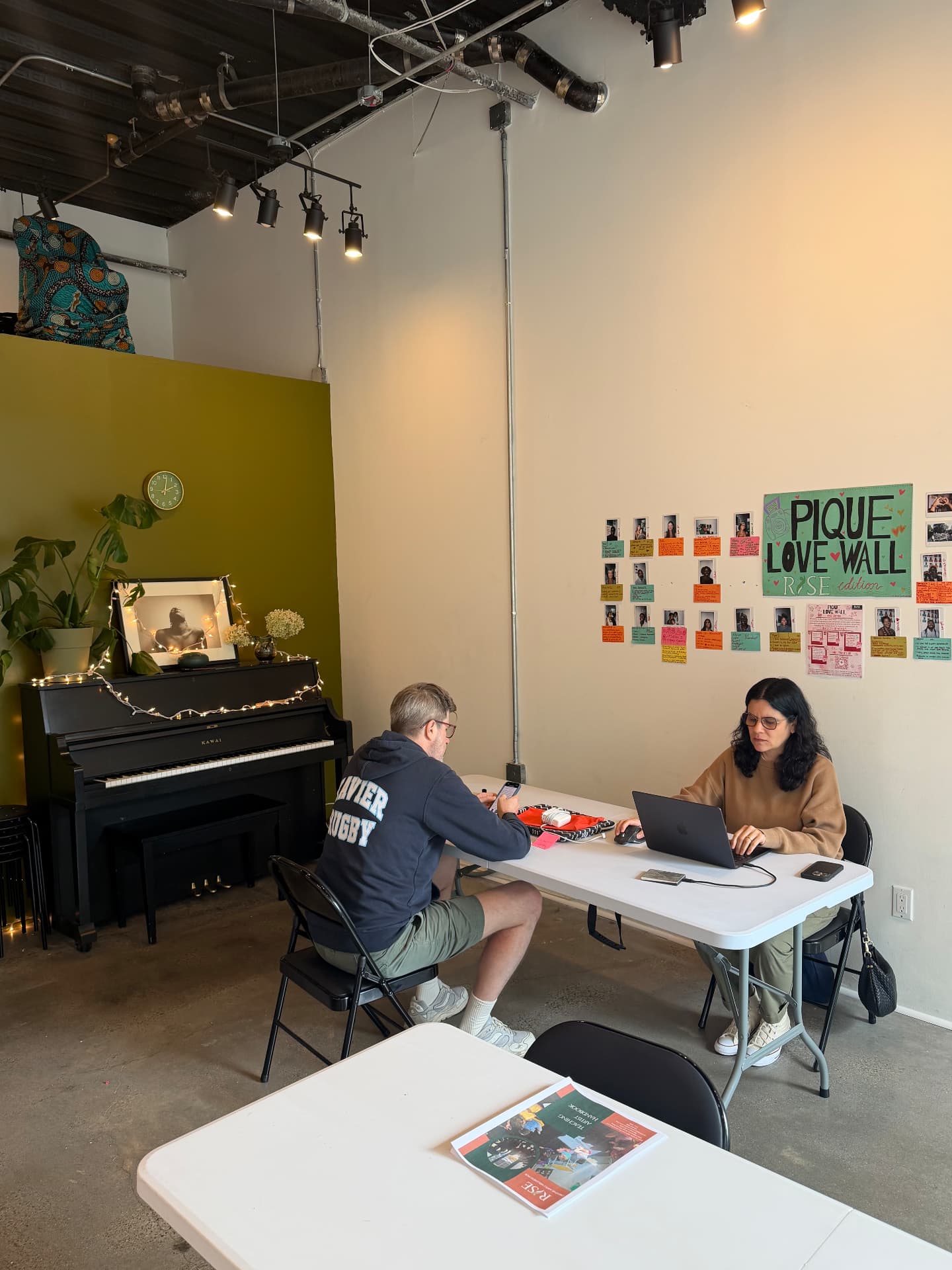 Two people working at a shared table at RISE, with the community wall behind
